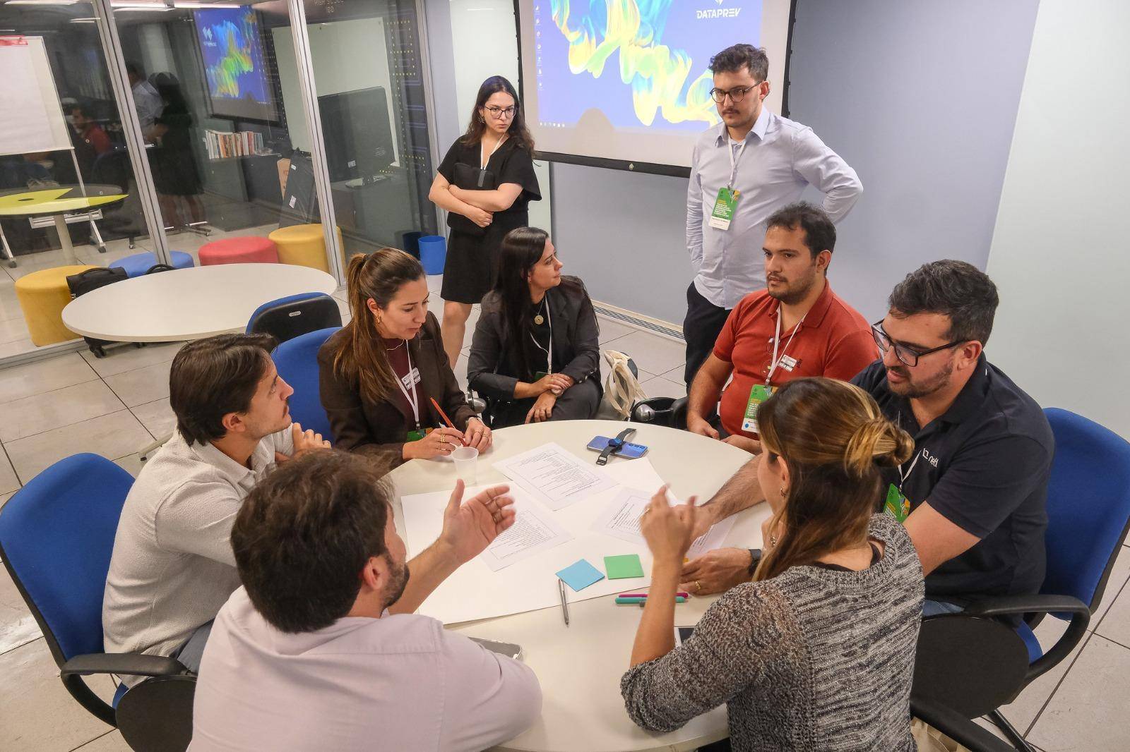 Nessa imagem podemos ver o grupo de trabalho ao redor de uma mesa. Existem sete pessoas sentadas ao redor da mesa, enquanto outras duas observam o grupo de pé.